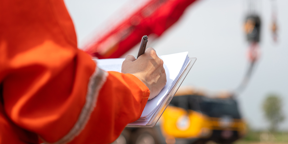 A safety inspector in hi-vis workwear filling out a lifting equipment inspection checklist on a clipboard with a crane in the background.