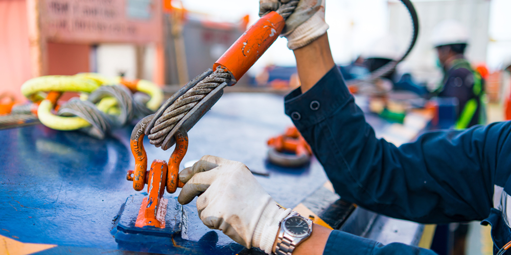 A close-up of an industrial rigger, wearing white safety gloves and a watch, conducting an inspection and adjustment of a heavy-duty orange steel bow shackle connected to a wire rope sling socket eye, on a blue industrial platform.